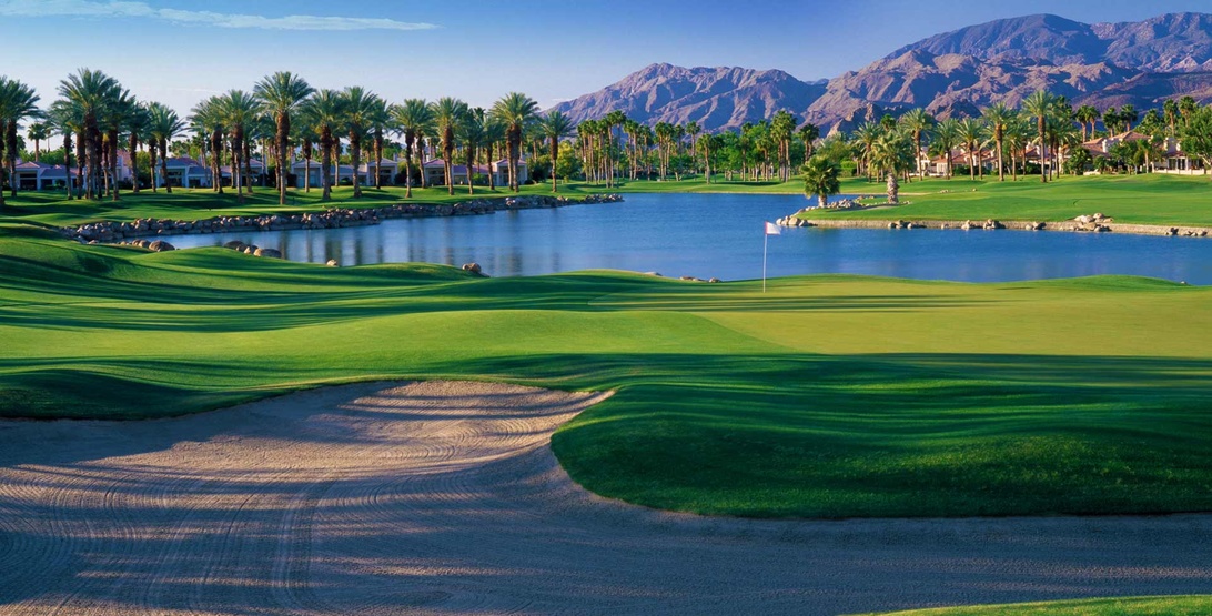En profesjonelt fotografert golfbane i ørkenlandskap med dramatiske vannhindere, palmer og fjellbakgrunner. Bildet viser det ferdige banedesignet med skulpturerte fairwayer, sandbunkere og et vannelement, men inneholder ingen tegn til anleggsarbeid.